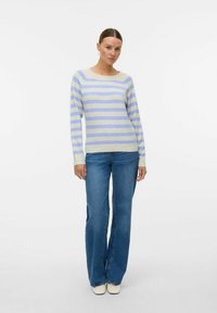 Maglione grigio chiaro con righe blu, scollatura rotonda, maniche lunghe. Abbinato a jeans in denim blu a gamba larga e scarpe bianche.