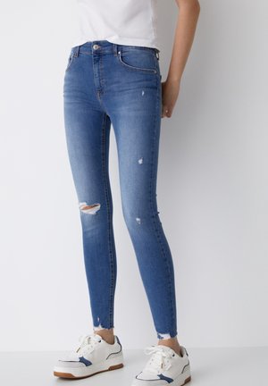 Person wearing slim-fit blue ripped jeans, white sneakers with blue details, and a white top, standing against a white background.