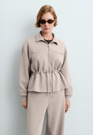 Femme portant une veste beige à fermeture éclair avec cordon à la taille et un pantalon assorti, debout devant un fond bleu pâle, portant des lunettes de soleil foncées.
