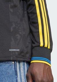 Black athletic jersey with yellow stripes on sleeves, AEROREADY logo, textured fabric featuring faint patterns, and contrasting blue cuff accents.