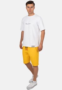 Witte T-shirt met korte mouwen en zwart logo, gecombineerd met gele shorts met vo pockets en een trekkoord. Witte sneakers met rode accenten.