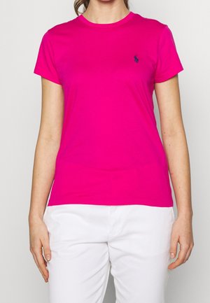 Femme portant un t-shirt rose vif à manches courtes avec un petit logo sur la poitrine et un pantalon blanc, debout devant un fond uni.