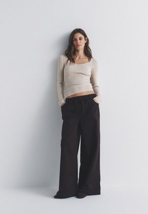 Noor naine seisab vastu seina, kandes beežikat pikkade varrukatega kitsast tippu ja laia jalaga lahtiseid musti pükse, käed taskutes.