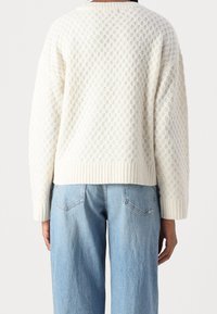 Cremfarbener Pullover mit einem strukturierten Blasenmuster, geripptem Saum und Bündchen, getragen über hellblauen Jeans. Der Rückenausschnitt ist leicht abgerundet.