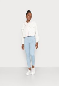 Casaco de ganga branco, top rosa e jeans skinny azul claro; modelo usa ténis brancos; pose em pé sobre um fundo simples.