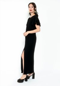 Vestido maxi negro con mangas acampanadas, abertura lateral y textura suave. Combinado con zapatos de plataforma negros, con una correa alrededor del tobillo.
