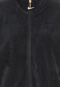 Černá kordurová mikina na zip s vysokým límcem, zlatým zipem a malým černým logem Nike v levém horním rohu.