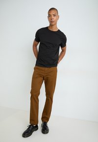 Black short-sleeve t-shirt paired with brown slim-fit trousers and black lace-up boots with yellow stitching, set against a light backdrop.
