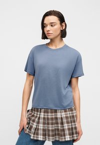 Νεαρός με κοντά σκούρα μαλλιά, φορώντας μπλε κοντομάνικο πουκάμισο πάνω από καρό καφέ πλισέ φούστα και μπλε τζιν.