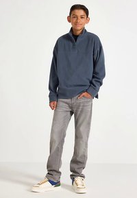 Modrý fleecový svetr na půl zip se šedými džínami a bílými teniskami s modrými a zelenými akcenty. Volný střih s dlouhými rukávy a žebrovanými manžetami.