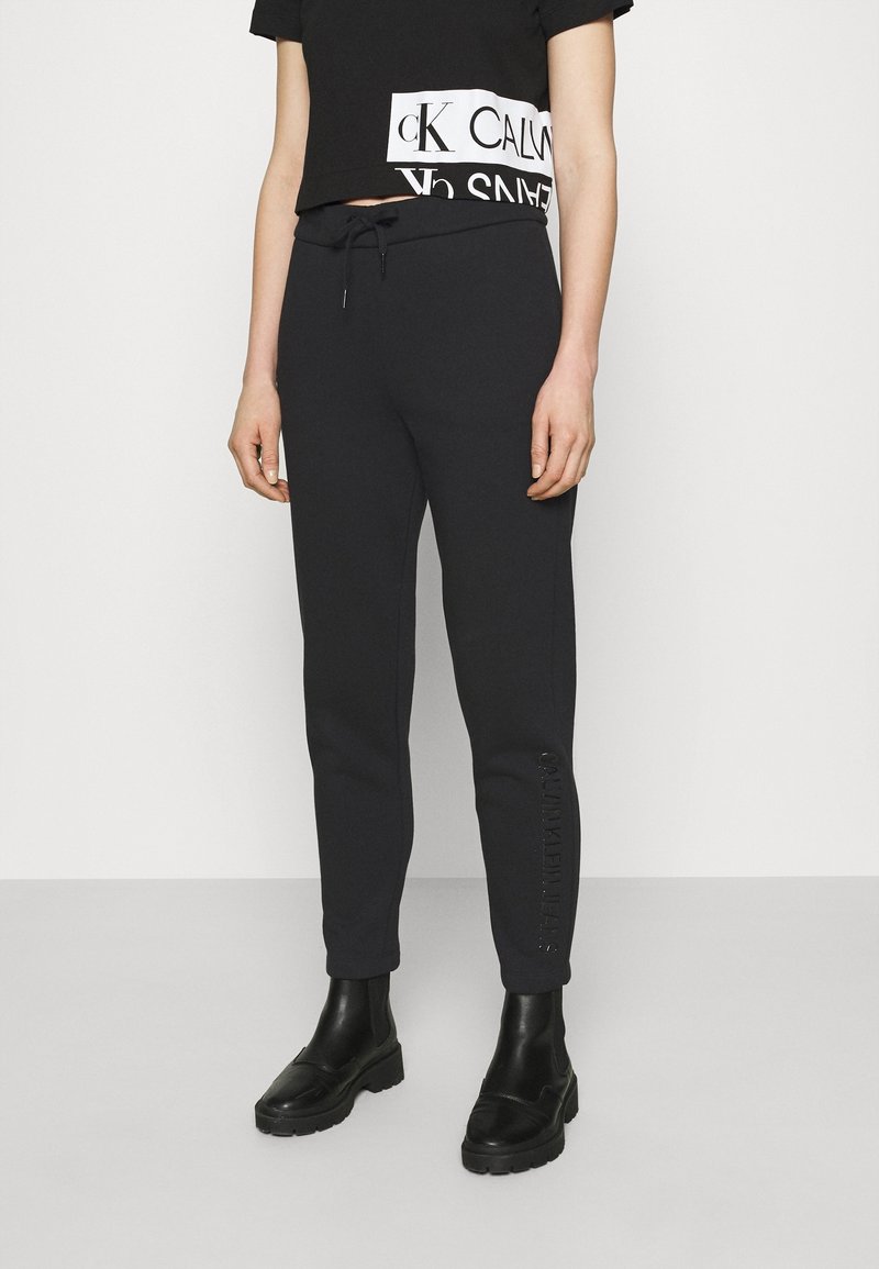 Svarta träningsoveraller i mjukt material med tapered design, utrustade med elastisk midja och en logotypdetalj längs vänster ben.