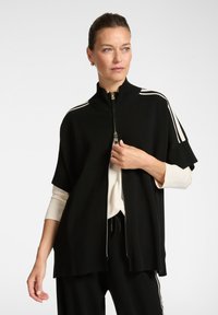 Czarna kurtka zapinana na zamek z wysokim kołnierzem, z białymi paskami na ramionach. Miękki dzianinowy materiał, luźny krój i ściągacz na dole.
