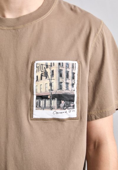 Pruun puuvillane T-särk, millel on tekstuuriga plaastriga linna stseeniga illustreering, valge äärega, paigutatud paremale rinna küljele.
