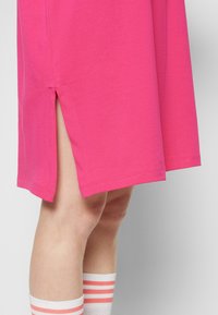 Fuchsia bomullsklänning med sidoklyfta. Sömmen är rak och tygtexturen är slät. Vita strumpor med rosa ränder synliga nedanför.