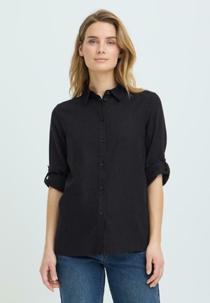 Mujer con cabello rubio hasta los hombros, vestida con una camisa negra abotonada con mangas remangadas y jeans azules, de pie contra un fondo liso.