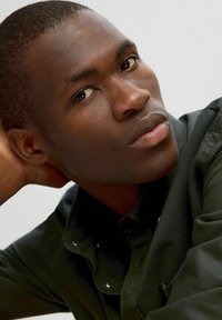 Primer plano de una persona con cabello corto y oscuro, vistiendo una camisa verde oscuro. La piel tiene una textura suave, acentuada por una iluminación uniforme.