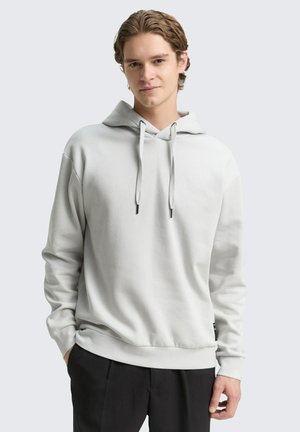 Junger Mann mit kurzen braunen Haaren, der einen schlichten hellgrauen Hoodie und schwarze Hose trägt, steht mit einer Hand in der Tasche.