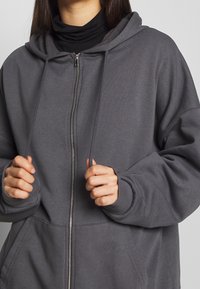 Sudadera gris con cremallera y corte relajado, con cordones y puños acanalados. El material parece suave y texturizado; cuenta con un bolsillo frontal.