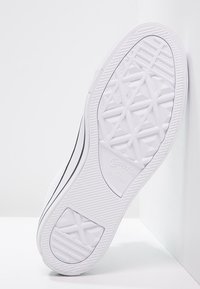 Sole bílé tenisky Converse All Star ukazující texturovaný vzor podrážky a černé boční pruhy, umístěná vzpřímeně na bílém povrchu.