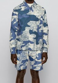 Mies, joka pukeutuu sinivalkoraidalliseen pitkähihaiseen nappipaitaan ja samaan kuosiin shortsit, seisoo yksinkertaista taustaa vasten.