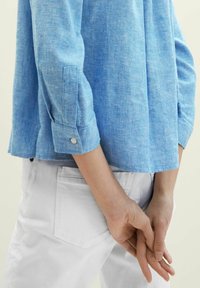 Chemise en lin bleu clair à manches longues, poignets boutonnés, associée à un pantalon blanc. Le tissu présente une texture subtile, créant un look décontracté.