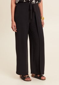 Zwarte wijde broek van lichtgewicht stof. Heeft een trekkoord in de taille en zijsplitten. Gecombineerd met zwarte instapsandalen.