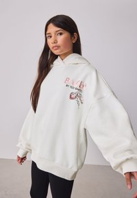 Balta didelio dydžio džemperis su gobtuvu, pagamintas iš minkštos medžiagos, su gobtuvu, ribotais rankogaliais ir siuvinėtu grafiniu užrašu priekyje su gėlių akcentais.