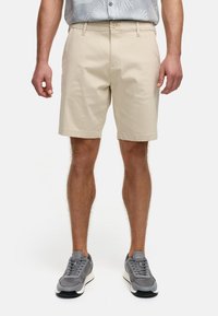 Beige knælange shorts lavet af bomuld med flad front, sidelommer og knaplukning, parret med grå sportsko.