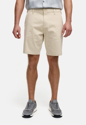 Shorts beige arrivés au genou en coton, avec un devant plat, des poches latérales et une fermeture à bouton, associés à des chaussures de sport grises.