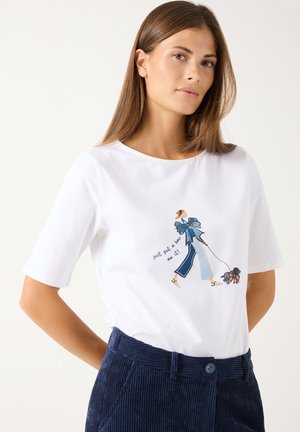 Frau trägt ein weißes T-Shirt mit einer Illustration einer gehenden Figur und einem Hund sowie dem Text "just put a bow on it,", kombiniert mit blauen Cordhosen.