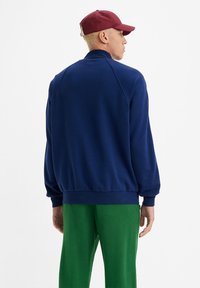 Mørkeblå sweatshirt med falsk krage, ribbede mansjetter og søm, kombinert med grønne bukser. Stoffet ser mykt ut med en jevn tekstur.