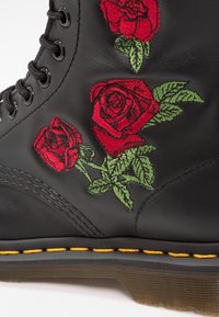 Bottes en cuir noir ornées de roses rouges brodées et de feuilles vertes, avec des lacets noirs et une semelle en caoutchouc cousue de fil jaune.