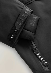 Schwarzer Stoff mit matter Oberfläche; ausgestattet mit einem Reißverschluss und einem Klettpatch mit "BAKER"-Branding. Glatte Textur, elegantes Design, funktionale Akzente.