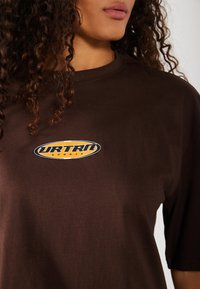 T-shirt en coton marron avec un logo circulaire orange et noir portant les mots "URTRU SPORTS". Le t-shirt a une coupe décontractée avec des manches courtes.