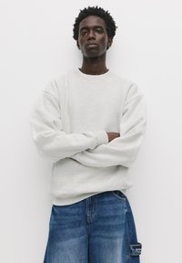 Hanorac ușor gri, cu gât rotund și mâneci ample, purtat împreună cu pantaloni din denim albastră, echipați cu un buzunar lateral. Textură netedă, design casual.