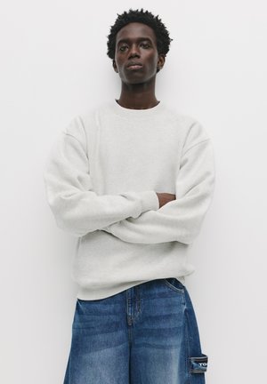 Light gray crew-neck sweatshirt with loose sleeves, paired with blue denim pants featuring a side pocket. Smooth texture, casual design.