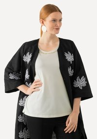 Sort linned kimonojakke med hvid bladbroderi, båret over en lys cremefarvet top. Har en rund halsudskæring og sorte bukser.
