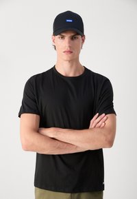 Black T-shirt with short sleeves and crew neckline. Paired with a black cap featuring a blue "HUGO" logo patch on the front.