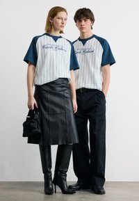 Νεαρή γυναίκα και άνδρας στέκονται όρθιοι, και οι δύο φοράνε ταιριαστά λευκά και ναυτικό μπλε κοντομάνικα πουκάμισα· η γυναίκα με δερμάτινη φούστα και μπότες, ο άνδρας με μαύρο παντελόνι.