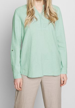 Femme portant un chemisier vert clair à manches longues et un pantalon beige, debout devant un fond gris clair uni.