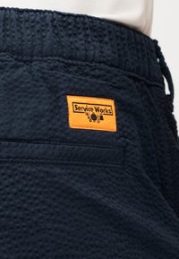 Close-up van een textuurrijke navy blauwe shortzak met daarboven een oranje label met 'Service Works' erop vastgenaaid.