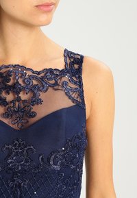 Närbild av en marinblå klänning med genomskinlig spets och paljettbroderi på den övre bröstet och axelbanden, bärs av en kvinna.