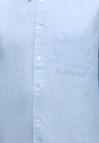 Chemise bleu clair à boutons avec une poche poitrine sur le côté gauche, montrant la texture du tissu et des boutons blancs sur le devant.