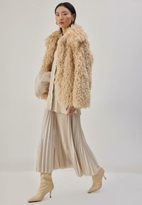 Beżowy płaszcz z futerka syntetycznego o puszystej teksturze, zestawiony z kremową plisowaną spódnicą i pasującymi szpilkami, z satynową torebką w dłoni. Subtelny połysk na całej powierzchni.