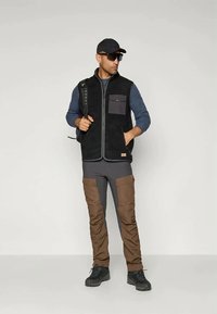 Gilet en polaire noir avec des accents gris, associé à une chemise à manches longues gris foncé et un pantalon cargo marron. Complété par une casquette et des lunettes de soleil.