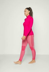 Top mulat, cu mânecă lungă, în roz aprins, asociat cu colanți roz care au accente stripe. Textură netedă și design sportiv.