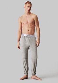 Szürke, elasztikus mandzsettával és fehér derékrésszel ellátott melegítőnadrág, amelyen a "Calvin Klein" logó látható, modell ing nélkül.