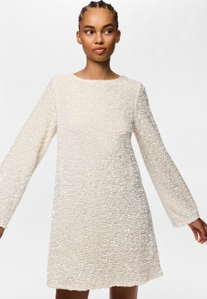 Mujer que lleva un vestido blanco texturizado de manga larga, de pie frente a un fondo blanco liso con los brazos ligeramente extendidos.