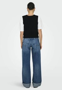 Schwarze ärmellose Strickweste über einem weißen T-Shirt getragen, kombiniert mit weit geschnittenen blauen Jeans mit umgeschlagenen Säumen und standardmäßigen Gesäßtaschen.