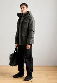 Giacca isolante verde oliva con molte tasche, indossata su un maglione grigio. Pantaloni neri e scarpe nere slip-on dalla trama, mentre si porta una borsa nera.